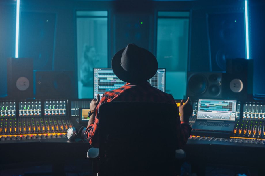 Producer, Audio Engineer Uses Control Desk for Recording New Album Track in Music Record Studio, in the Soundproof Room Musician, Artist, Performer Sings a Song from New Album. Back View